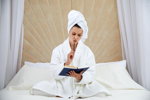 Young woman sits on bed wearing bathrobe, making notes in diary. Blogger creats publication for blog in notebook