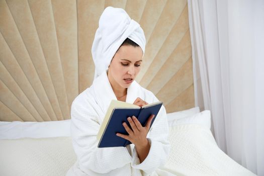 Young woman sits on bed wearing bathrobe writing notes in diary. Blogger creats publication for blog in notebook