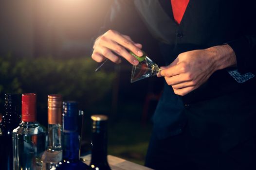 Professional bartender preparing fresh lime lemonade cocktail in drinking wine glass with ice at night bar clubbing counter. Occupation and people lifestyles concept. Outdoor background