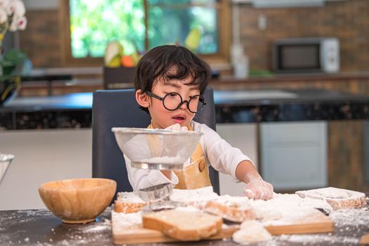 Cute little Asian boy sifting dough flour with sifter sieve colander in home kitchen for prepare to baking bakery and cake. Thai kids playing with flour as chef funny. People child playing