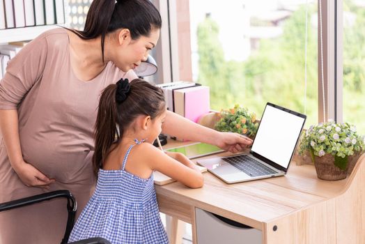 Cute daughter and her pregnant mother using white blank screen laptop at home. Parenthood and technology. Health and medical. People lifestyles and family concept. Copy space