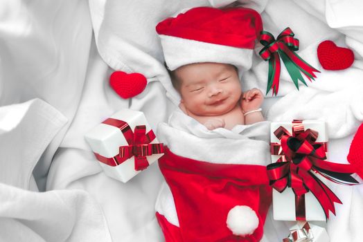 Sleeping newborn baby face in Christmas hat with gift box from Santa Claus and yarn heart on white soft towel. Cute Infant lifestyle and innocent happy baby lying in cold snow season. New year winter