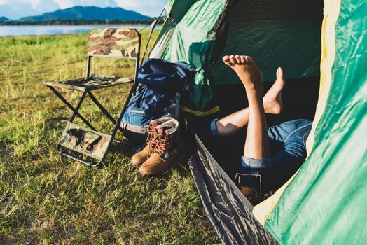 Close up of woman legs relaxing in camping tent with mountain lake and meadow and grass field background. Lifestyles and People concept. Camping and picnec theme. Green natural and summer travel theme