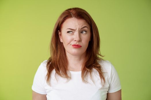 Perplexed doubtful unsure redhead wife smirking unsatisfied frowning look aside displeased disappointed hesitating pondering strange advice thoughtfully stare away stand green background