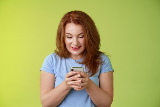 Intrigued excited tempting redhead middle-aged woman shopping online smartphone hold mobile phone look pleased entertained gadget screen smiling delighted playing game found perfect app