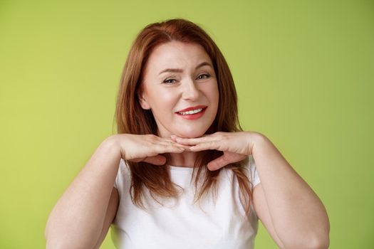 Looking good. Happy cheerful redhead middle-aged 50s woman smiling delighted hold hands under chin accept flaws blemished like own skin condition apply aging creme cosmetics green background