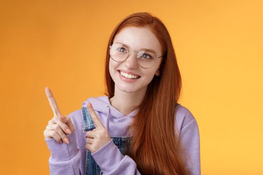 Carefree charismatic happy silly young cute redhead girl freckles blue eyes wearing glasses enjoy contemplating entertaining exhibition pointing looking left indicating, orange background