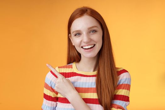 Friendly outgoing good-looking ginger girl university student discussing lecture classmate smiling laughing pointing upper left corner questioned curious know details standing orange background