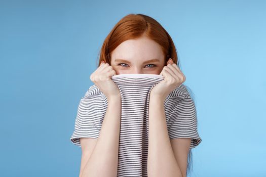 Silly flirty amused attractive playful redhead girlfriend hiding face pulling t-shirt head squinting devious mysteriously giggle laughing hope disguise pranking friend standing blue background