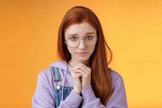Silly guilty young shy redhead girlfriend asking forgiveness supplicating lower head look from under forehead frowning begging apology favour standing insecure sad pleading help, orange background