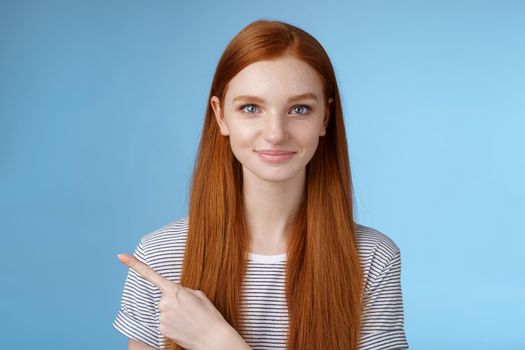 Attractive modest good-looking redhead girlfriend acting mature friendly smiling pointing left index finger giving direction show way bathroom standing blue background joyful kind grin