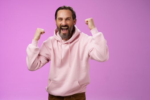 Cheerful supportive manly mature adult bearded guy fan yelling raising clenched fists triumphing team scored goal celebrating standing pleased shouting achieving success, posing purple background