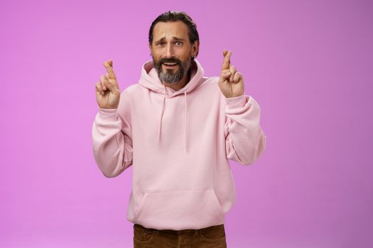 Nervous unsure hopeful handsome bearded 40s man in pink stylish hoodie cringing worried cross figers good luck make wish hope not gonna lose job supplicating praying dream come true