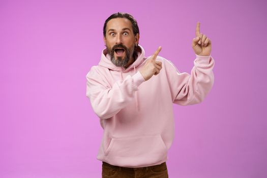 Impressed shocked exctied cool hipster mature 50s man bearded grey hair in pink hoodie retelling incredible story pointing up index fingers widen eyes surprised thrilled awesome advertisement