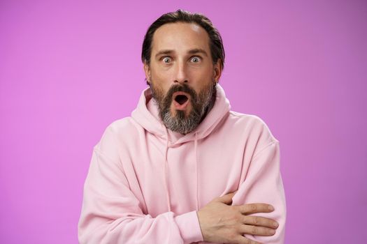 Shocked impressed gossiping caucasian bearded adult 40s man grey hair in pink hoodie gasping fascinated fold lips wow widen eyes astonished hearing interesting thrilling story, purple background