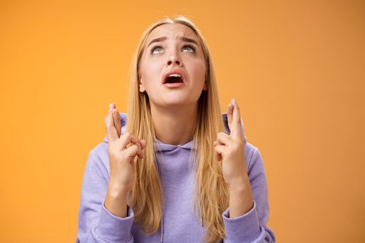 Worried nervous cute blond woman in hoodie begging god help cross fingers good luck supplicating look up skies wanna win anticipating good news anxiously awaiting, orange background