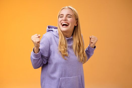 Cute happy triumphing blond european girl in hoodie yelling yes accomplish dream lifetime clench fists happily dancing celebrating victory success achieve goal, standing orange background