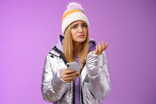 Questioned complicated cute blond girlfriend receive strange message look perplexed confused raising hand shrugging lift eyebrow cannot understand meaning holding smartphone, purple background