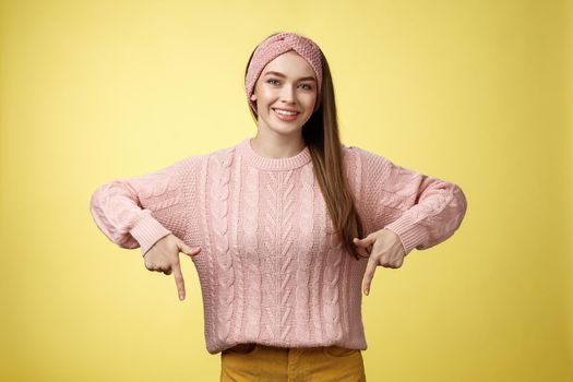 Tender young 20s european girl in sweater smiling broadly finally take-off braces pointing down promoting skillful dentist professional. Female student standing happy positive over yellow wall