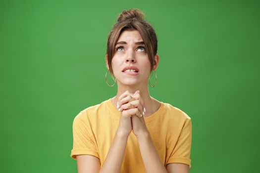 Intense nervous and faithful good-looking young woman clenching hands together over chest looking up and biting lower lip while praying god help, standing hopeful over green background