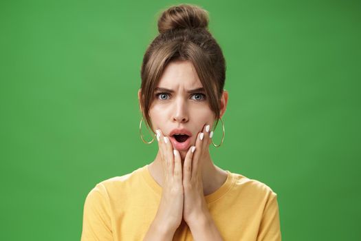 Shocked woman reacting to pimple looking disturbed and displeased in mirror holding hands pressed to cheeks open mouth and frowning being disappointed and upset over green background