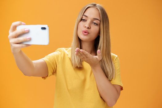 Stylish glamourous female fashion blogger ending recording video via smartphone by sending air kiss at camera, taking selfie with sensual and confident gaze at screen posing over orange wall