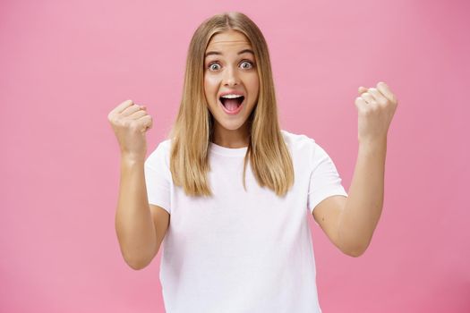Excited cheerful and optimistic charming woman with fair hair in white t-shirt raising fists in victory and triumph yelling yes in success and amazement standing supportive against pink background