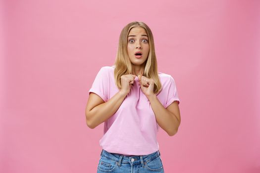 Woman gasping from fright being scared by someone pressing clenched hands to chest bending backwards looking worried and terrified at camera being shocked with unexpected bang over pink wall