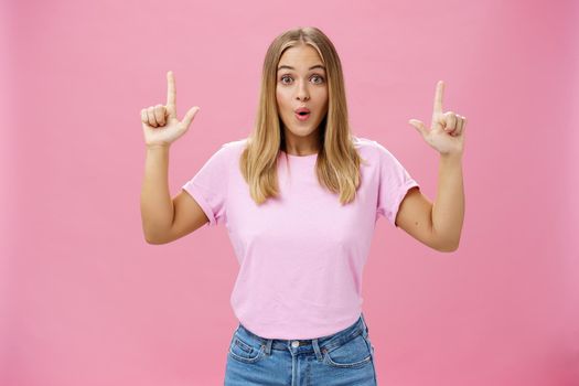 Amused and impressed attractive common european woman with fair hair and tanned skin raising index fingers pointing up amazed folding lips from interest and astonishment over pink background
