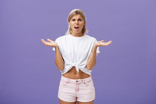 Perplexed and disappointment good-looking blonde female student in white t-shirt and pink shorts frowning shrugging with spread hands near shoulders saying what the hell over purple wall