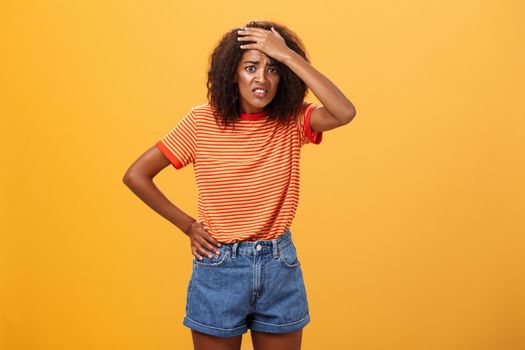 Shit I am in trouble. Concerned anxious cute dark-skinned woman in trendy t-shirt and shorts clenching teeth with guilty look frowning punching forehead having trouble or problem over orange wall