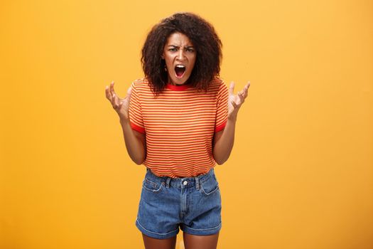 Pissed and outraged angry african american young female in stylish outfit raising palms gesturing and yelling losing temper arguing with cheating partner feeling fed up over orange background