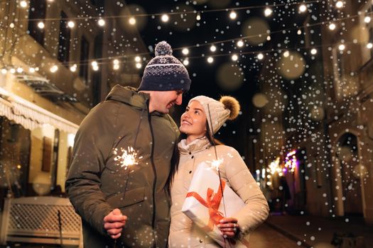 Lovers travelling to Europe at the height of Christmas holidays. Sweethearts in the centre of city. Romantic couple in love with gift box hugging and burning the sparklers.
