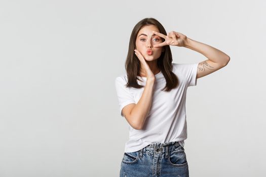 Beautiful young woman showing peace gesture and pouting silly, white background