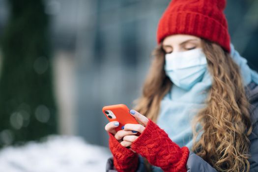 Close up portrait of beautiful Caucasian young curly hair female in medical mask texting on cellphone. Caucasian woman browsing on smartphone and looking away. Being online, social networks.