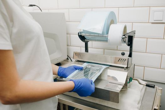 Dentist's assistant hands in gloves packing dental instrument set for autoclaving in a plastic bag using vacuum packing machine. Sterility and safety care concept.