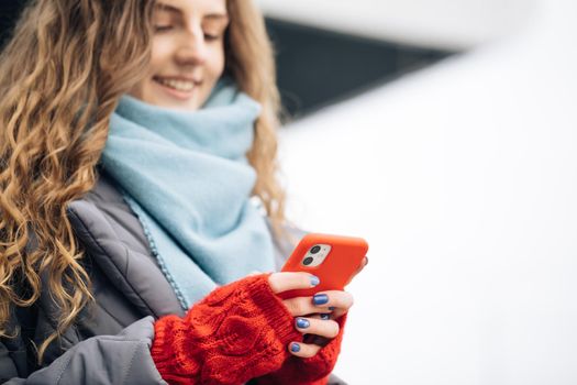 Portrait of cheerful Curly-haired Young female texting on smartphone standing on street in winter city on New Year. Female tapping on cellphone outdoors. Online shopping, buying new year's gifts.