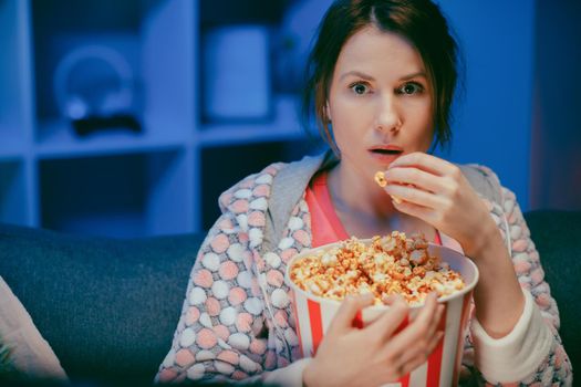 Portrait shot of the woman with popcorn sitting on the sofa watching something scary while eating popcorn and being afraid