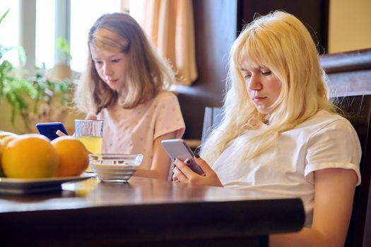 Children two girls sisters eating at home, looking at smartphones
