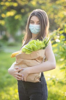 Vertical Portrait of Caucasian young pretty woman delivery worker in medical mask on the park carrying packet with fresh food.