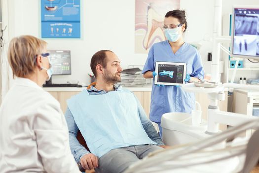 Dentist assistant showing teeth radiography on tablet pc computer screen to sick patient man at dental clinic office. Orthodontist discussing toothache treatment to prevent stomatology problems