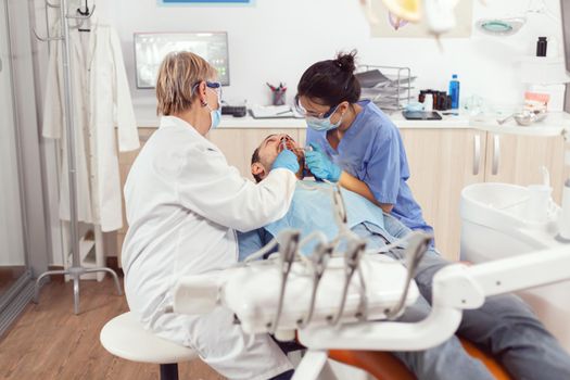 Senior woman dentist analyzing teeth examining sick patient while sitting on orthodontic chair. Doctor stomatologist and nurse cleaning mouth during dental examination in stomatology clinic