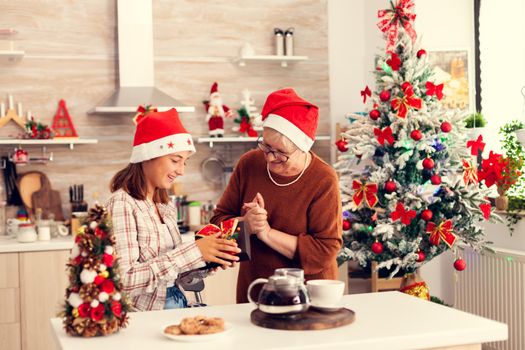 Cheerful granny and niece enjoing christmas celebration with presents in decorated home. Senior woman wearing santa hat surprising granddaughter with winter holiday present in home kitchen with xmas tree in the background.