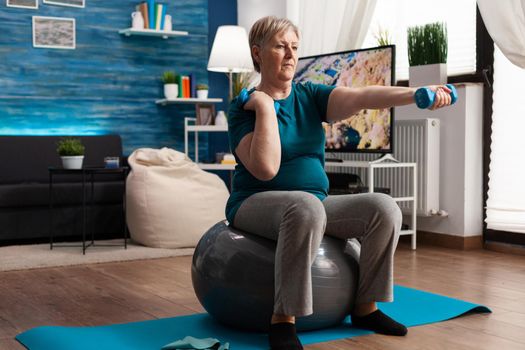 Athletic pensioner in sportswear watching online aerobic workout using tablet sitting on swiss ball