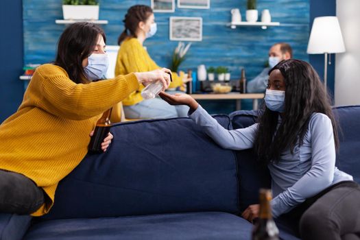 Group of multi ethnic using hand sanitizer to prevent coronavirus spread while spending time together in home living room sitting on couch wearing face mask. Conceptual image.