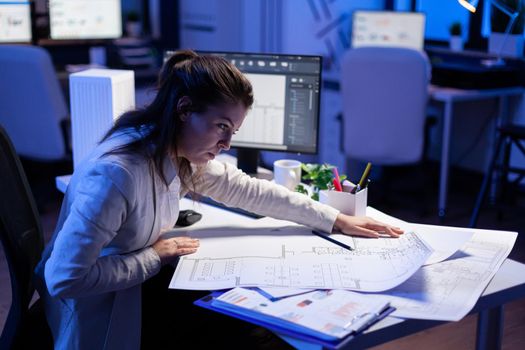 Overworked woman architect checking and matching blueprints sitting at office desk