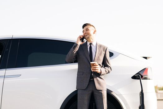 Businessman is talking on his Smart Phone while his electric car is charging on background. Side view of white luxury car. Environmentally conscious male charging electric vehicle