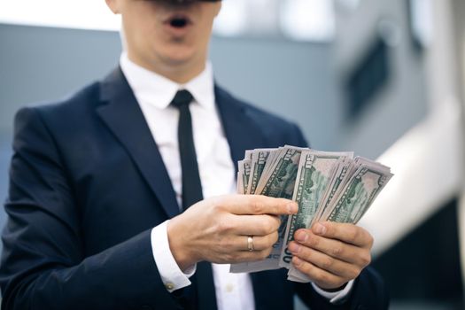Rich man wearing VR headset counting money and smiling. Young businessman standing with pack of dollars. Richness and success concept. New technology offers new 3D dimensions.