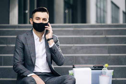Fired man in medical mask talking on phone with stuff in box. Cellphone conversation. Speaking on telephone. Handsome jobless office worker entrepreneur in formal suit sitting on stairs outdoors
