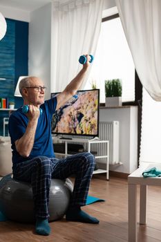 Retirement senior man sitting on aerobics swiss ball raising hand streching arm muscle